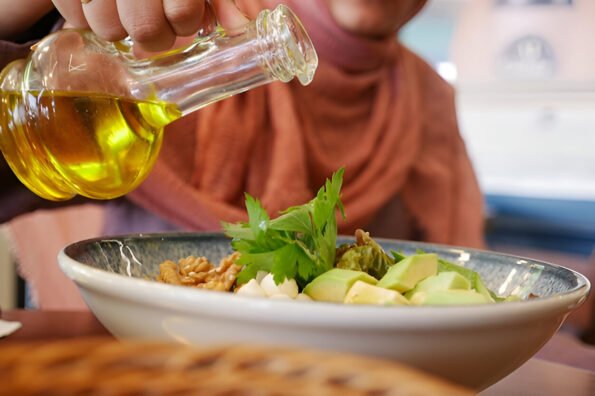 Person pouring olive oil from a glass bottle onto a fresh salad bowl with avocado, walnuts, and herbs